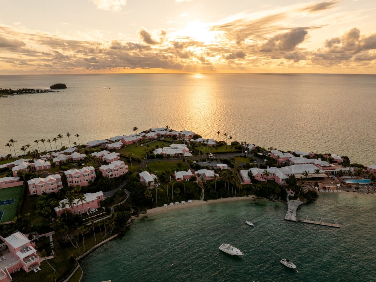 A tropical coastal resort with pink-roofed villas along a curved shoreline, anchored boats in calm turquoise water, and a golden sunset over the ocean.