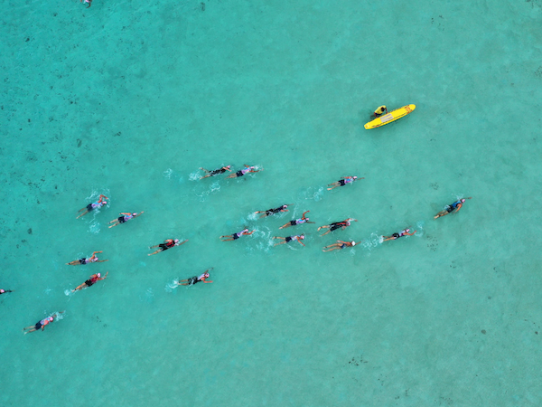 Aerial view of a turquoise ocean with a group of people paddleboarding or swimming, followed by a bright yellow boat nearby.