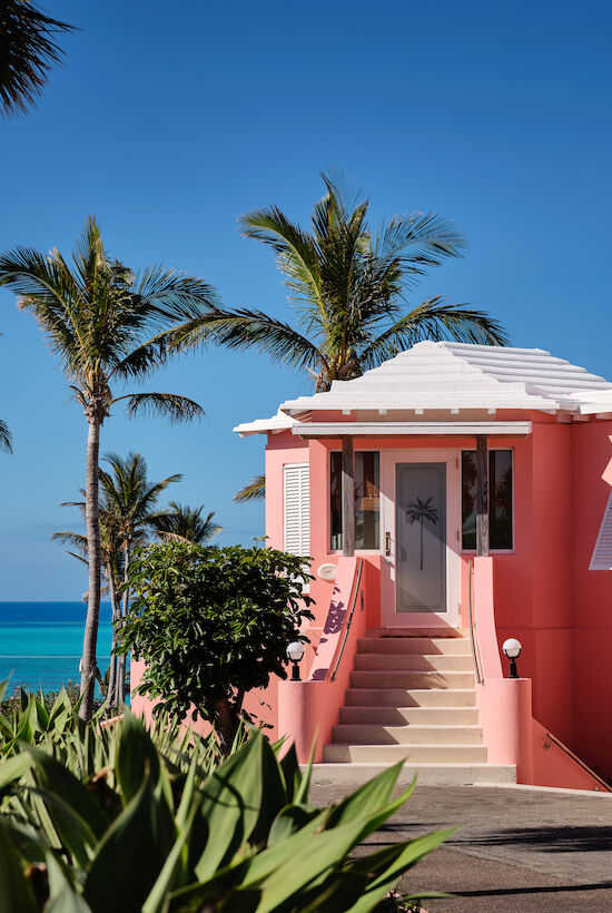 A pink house with white shutters and roof, surrounded by lush greenery and palm trees, overlooking a clear blue ocean under a bright sky.