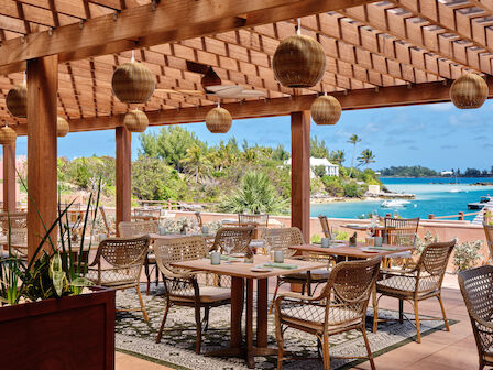 An outdoor dining area with wicker chairs, a wooden pergola, hanging lights, and a view of the sea and lush greenery.