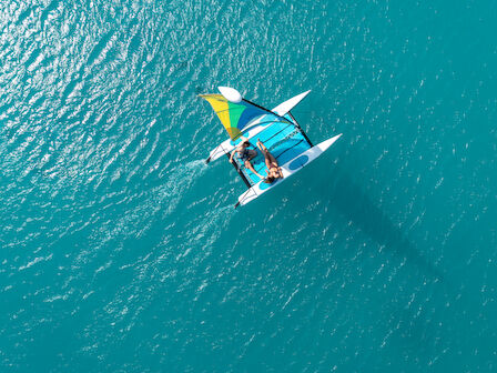 Aerial view of two people on a small sailboat with a colorful sail, navigating through clear blue water.
