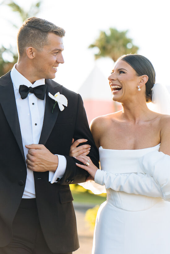 A couple in wedding attire walking arm in arm, both smiling joyfully with lush greenery in the background.