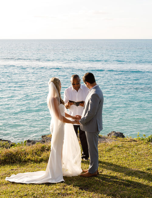 A couple is getting married on a grassy cliff by the ocean with an officiant.