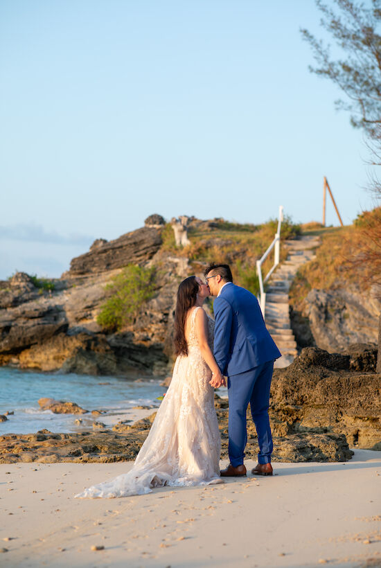 A couple in wedding attire shares a kiss on a sandy beach with rocky background and gentle waves under a clear sky.