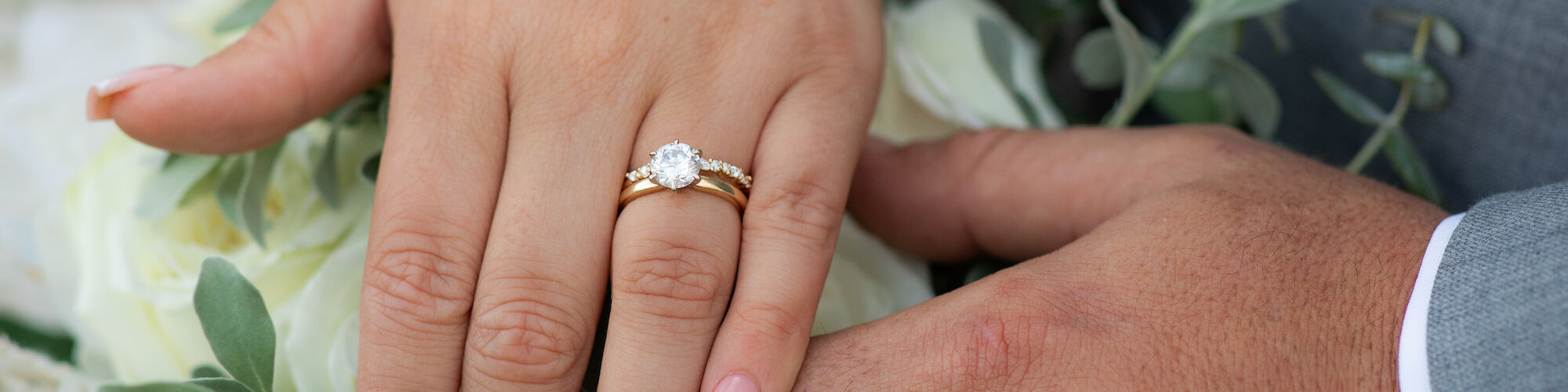 Two hands with wedding rings resting on a bouquet of white roses and greenery, with a close-up view capturing the intimate moment.