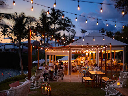 A cozy outdoor dining area with string lights, palm trees, and people enjoying a meal at dusk.