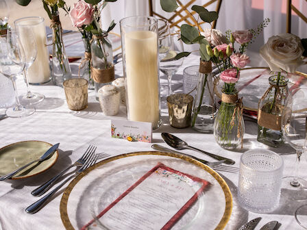 Elegant table setting with glass plates, floral arrangements, candles, and cutlery on a white tablecloth. A menu and name card included.