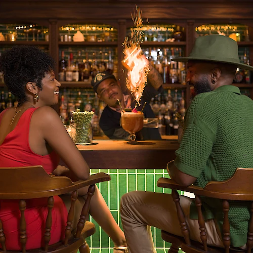 A couple sits at a bar while a bartender ignites a flaming drink in the background, creating a lively and engaging atmosphere.