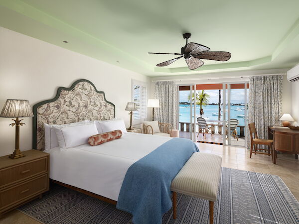 A cozy hotel room with a large bed, ceiling fan, and patio doors leading to a balcony with a beach view, featuring calm decor.