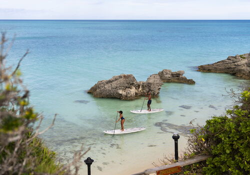 Two people are paddleboarding near rocks in clear, calm water, surrounded by greenery, under an overcast sky.