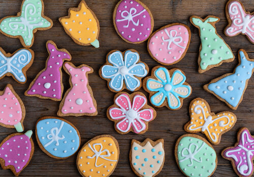 Colorful, decorated cookies shaped like butterflies, flowers, dresses, and eggs are arranged on a wooden surface.