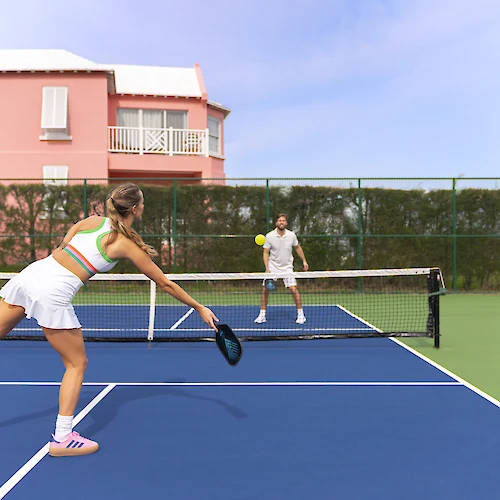 Two people are playing pickleball on a blue court, with a pink house and greenery in the background under a clear sky.