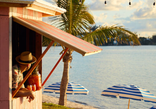 A small pink beach shack with a person inside, palm trees, string lights, and striped umbrellas by the water under a blue sky.