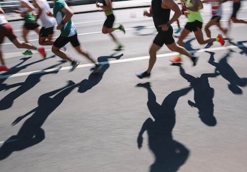 Runners are participating in a race, casting long shadows on the pavement as they move forward in the bright sunlight.