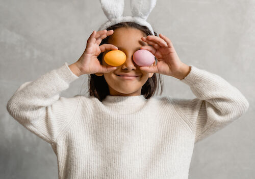 A child wearing bunny ears holds colored eggs in front of their eyes, smiling playfully against a neutral background.