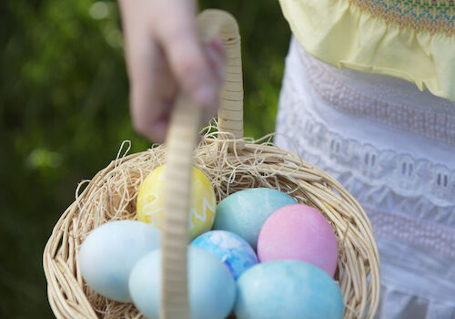 A person is holding a basket filled with pastel-colored Easter eggs, standing on grass.