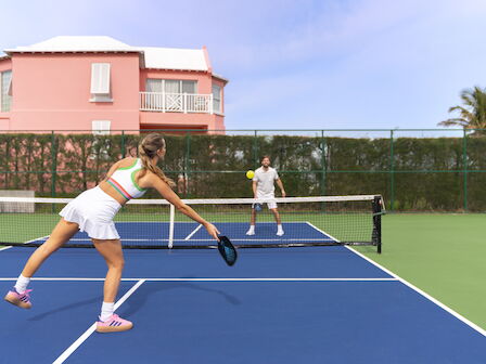 Two people are playing pickleball on a blue court near a pink house, surrounded by greenery.