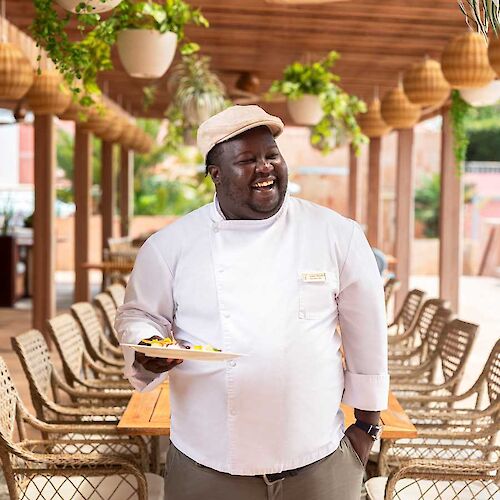 A person in a chef's uniform stands outside, holding a plate, smiling, with plants and wooden furniture around in a sunny setting.