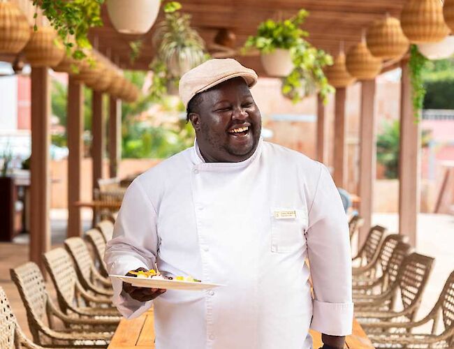 A person in a chef's uniform stands outside, holding a plate, smiling, with plants and wooden furniture around in a sunny setting.