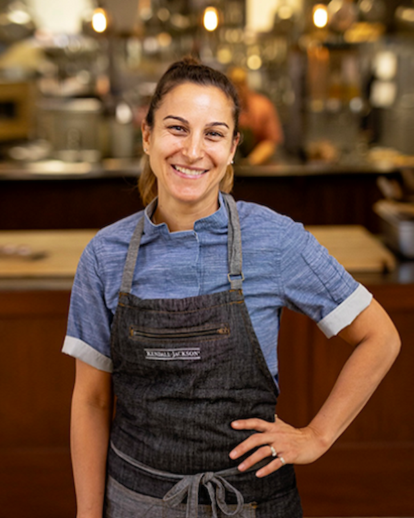 A person smiling in a commercial kitchen, wearing a blue shirt and apron, standing with hands on hips in a bustling restaurant setting.