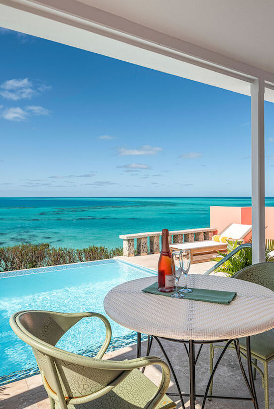 Patio by a pool with an ocean view, table with two glasses and a wine bottle, surrounded by lounge chairs, clear blue skies above.