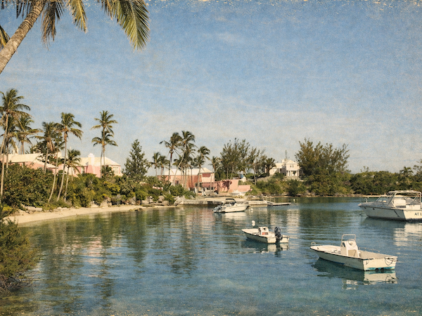 A tropical coastal scene with palm trees, calm blue water, boats anchored near a sandy shoreline and pastel houses in the background, during a sunny day.