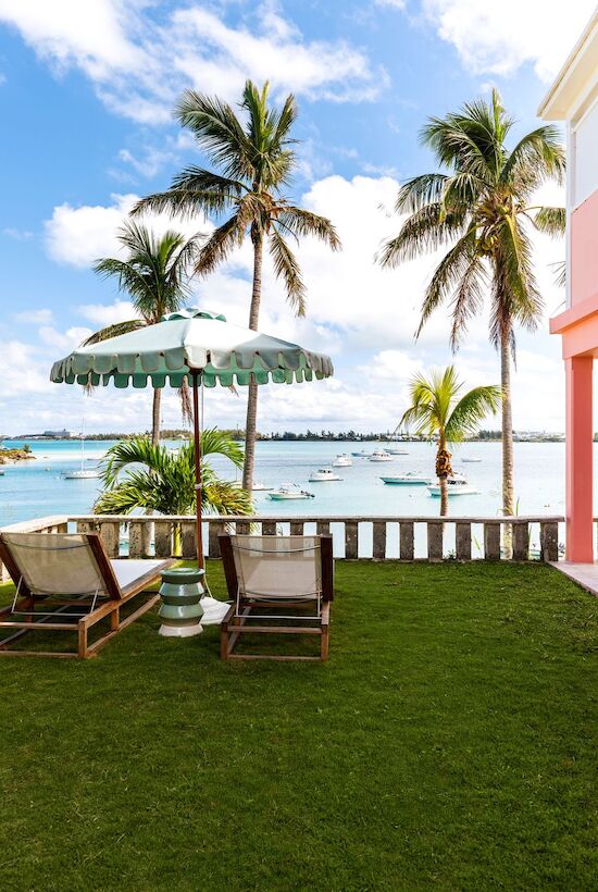 A tropical scene with a pink building, palm trees, two lounge chairs with an umbrella, and a view of the ocean and boats.