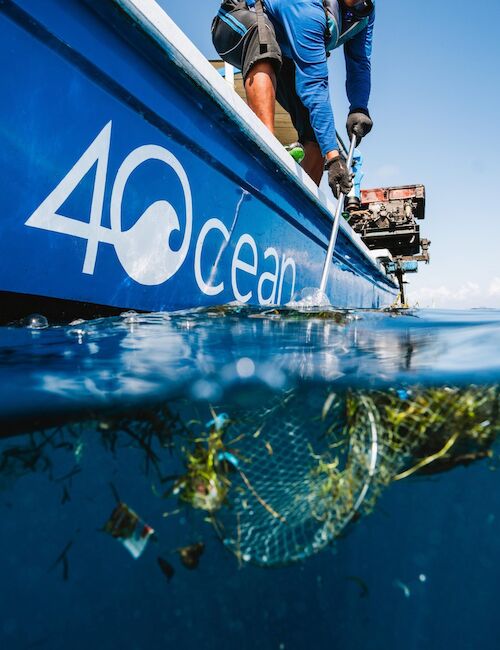 A blue boat with "4Ocean" logo, people cleaning trash from the ocean surface, net catching debris and seaweed.