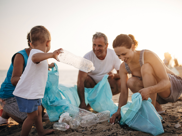A group of people, including a child, collect plastic bottles and trash on a beach, using blue bags under the setting sun.