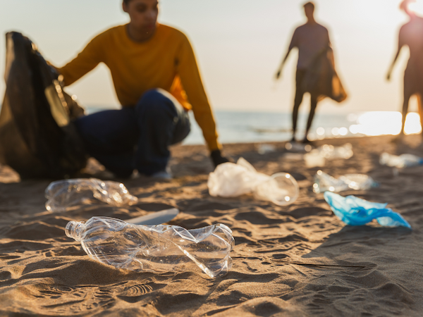 People cleaning up plastic waste on a sandy beach, with sunlight in the background.