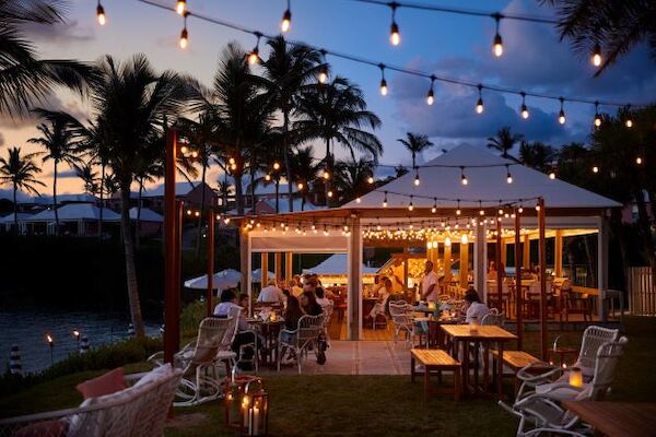Outdoor dining area with people, string lights overhead, and palm trees in the background at dusk, creating a cozy ambiance.