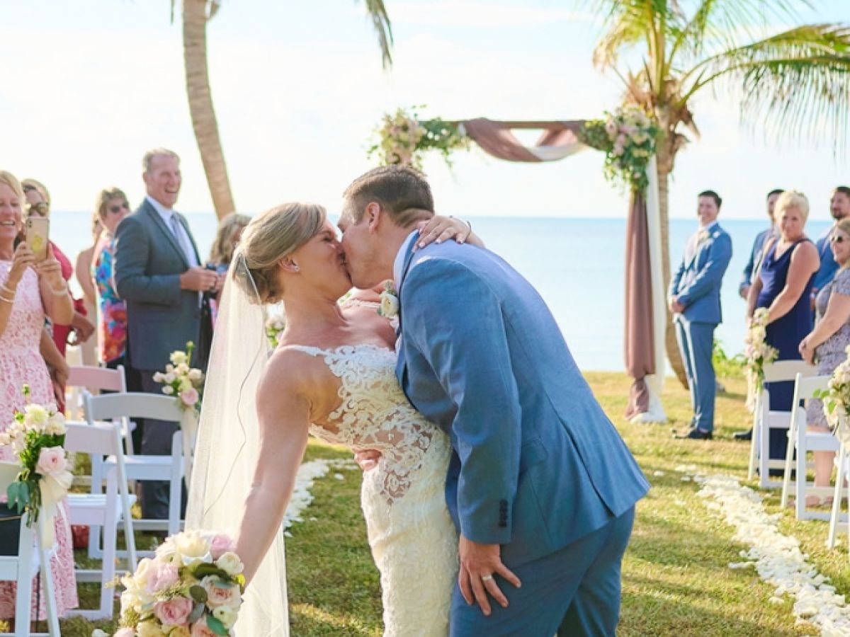 A newlywed couple shares a kiss under palm trees at an outdoor wedding ceremony surrounded by guests in a picturesque setting.