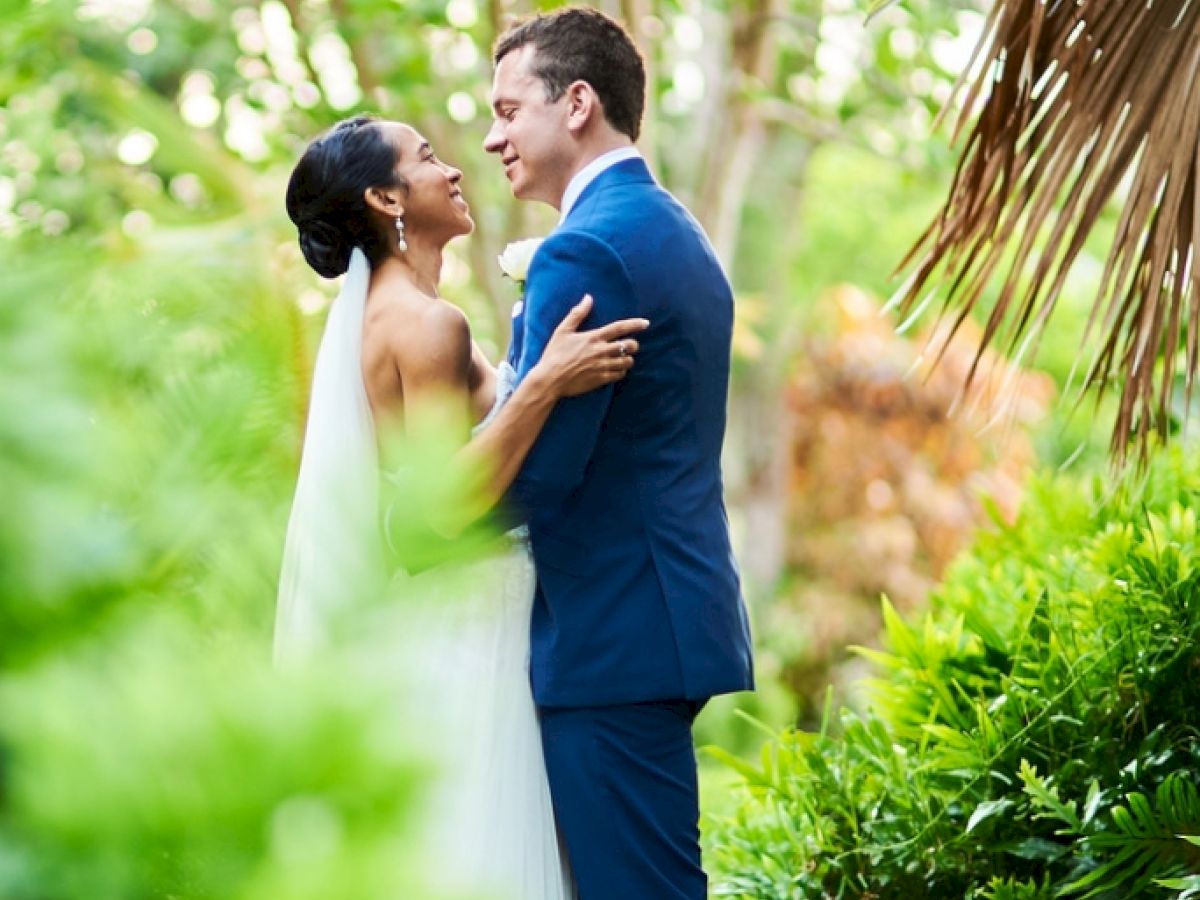 A bride and groom embrace lovingly in a lush, green garden setting, beneath a stone archway, sharing a special moment on their wedding day.