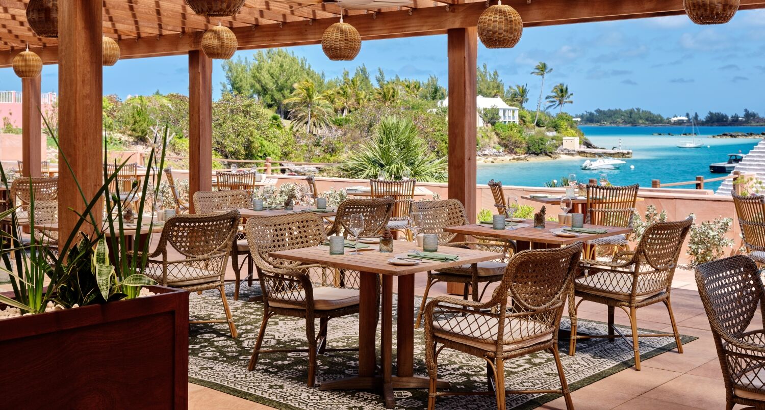 An outdoor seaside dining area with wicker chairs and round tables under a wooden pergola, overlooking a turquoise lagoon and boats, sunny and breezy.