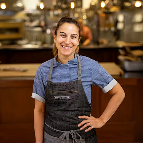 Person in a kitchen, wearing a denim shirt and dark apron, smiling with hands on hip, surrounded by culinary equipment and warm lighting.