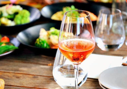 A table with a glass of rosé wine, plates of food, and other glasses, suggesting a casual dining setting.