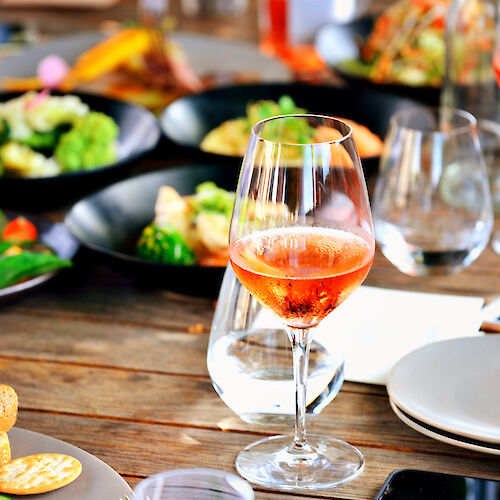 A table with a glass of rosé wine, plates of food, and other glasses, suggesting a casual dining setting.