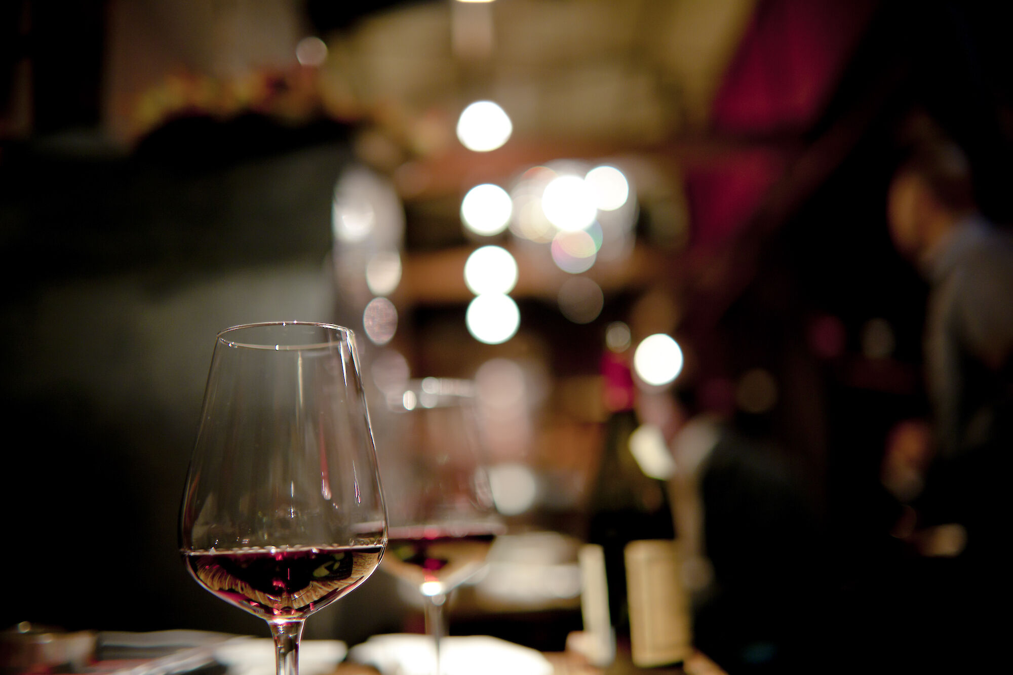 A close-up of wine glasses on a table in a dimly lit setting, with blurred lights creating a bokeh effect in the background.