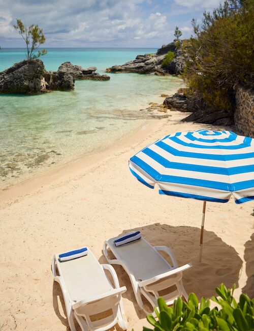 A sunny beach scene with clear turquoise water, sandy shore, rocky outcrops, and a blue-and-white striped umbrella over two white lounge chairs, ending with a calm horizon.