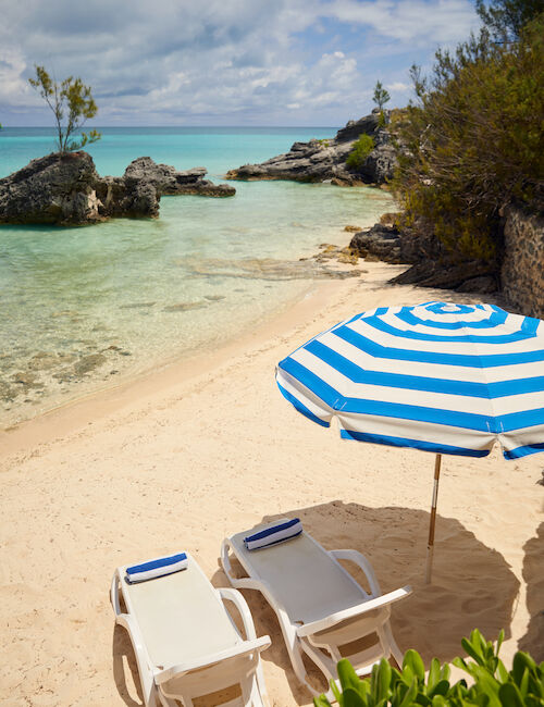 Two lounge chairs under a striped umbrella on a sandy beach with turquoise water and rock formations in the background.