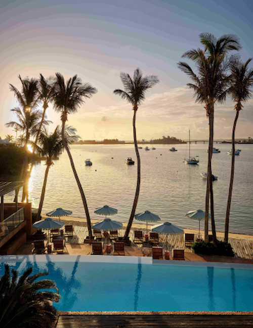 A serene waterside scene with a pool, palm trees, umbrellas, lounge chairs, and boats in the sunset-lit background.