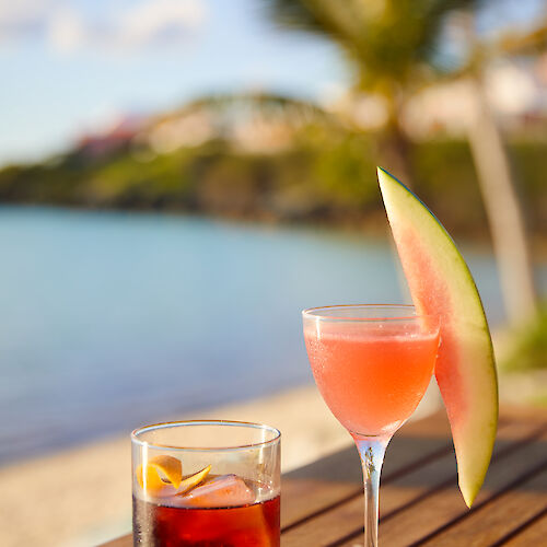 Two cocktails on a wooden table by the beach, one with an orange slice and the other garnished with a watermelon slice.