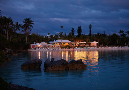 A beachside resort is illuminated at dusk, with palm trees and calm waters reflecting the lights.