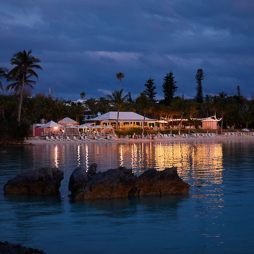 A beachside resort is illuminated at dusk, with palm trees and calm waters reflecting the lights.