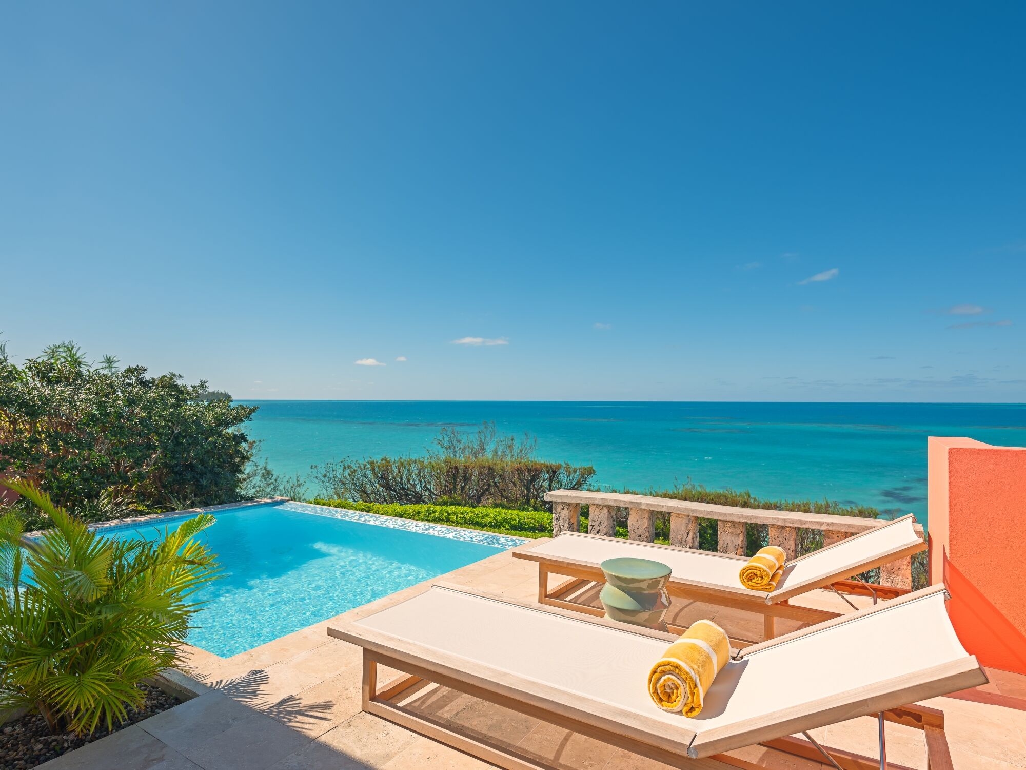 A sunny oceanfront scene with a turquoise pool, two sun loungers, rolled towels, and a clear blue sea meeting a bright sky.