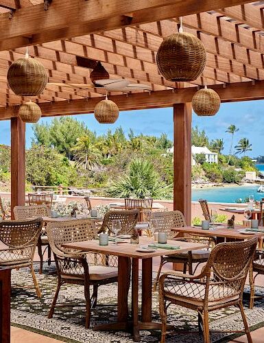 An outdoor seaside dining area with wooden tables and wicker chairs under a thatched roof, overlooking palm trees and turquoise water.