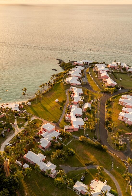 Aerial view of a coastal resort with a sunset over calm waters, white buildings clustered along winding roads and green lawns by the shore.