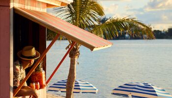 A beach scene with a wooden shack, a person, tropical drinks, palm trees, and blue-striped umbrellas near the water's edge.