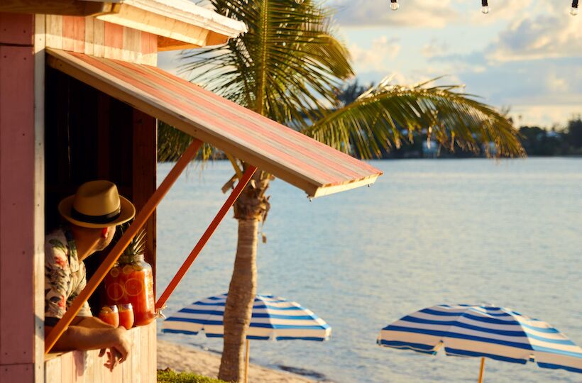 A beach scene with a wooden shack, a person, tropical drinks, palm trees, and blue-striped umbrellas near the water's edge.