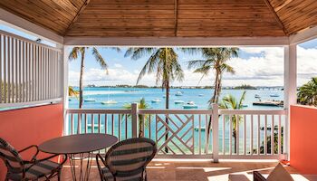 Covered patio with chairs and a table, overlooking a scenic view of palm trees, boats on blue water, and a clear sky beyond a railing.